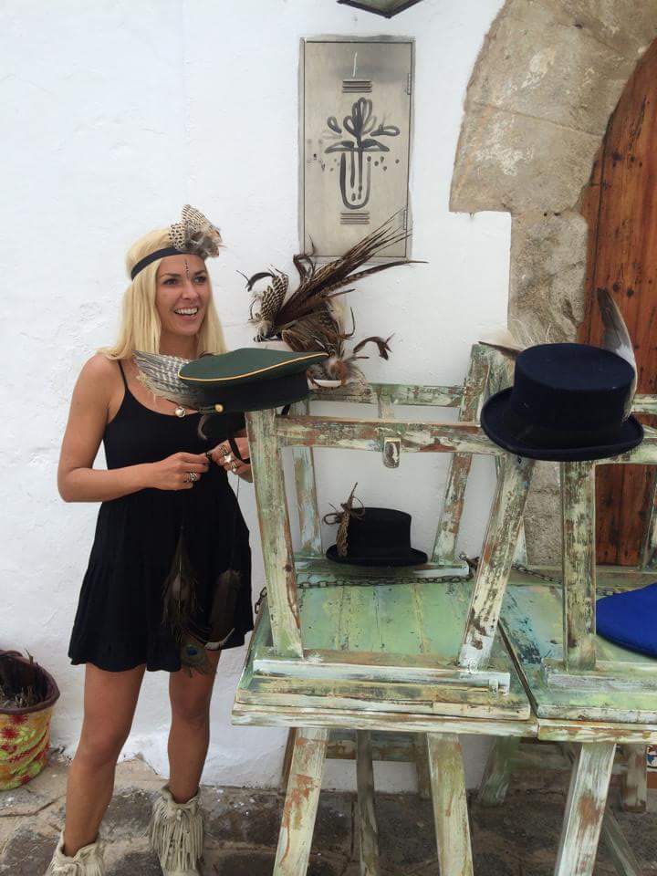 A blond lady smiling in front of some hats and feathered headdresses.